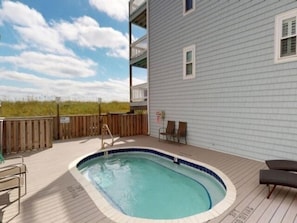 A swimming pool on a wooden deck with a view of the beach and a building with balconies.