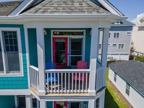 Een huis met twee verdiepingen, een overdekte veranda, een rode deur en een klein zitgedeelte buiten met stoelen.