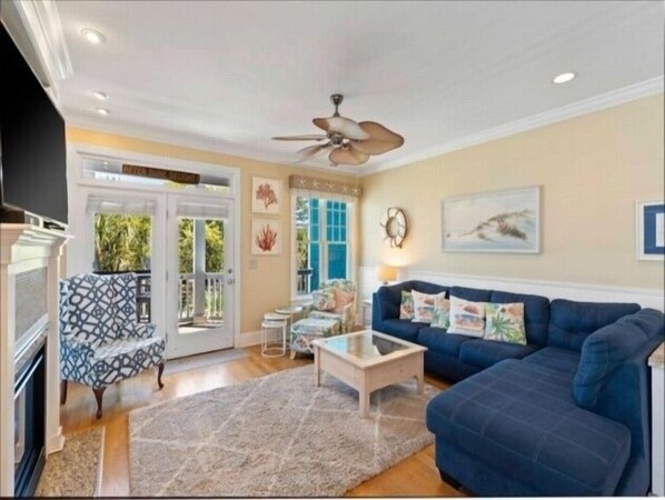 Een woonkamer met een blauwe bank, een salontafel en een plafondventilator. Er is een deur die leidt naar een balkon met uitzicht op groen.
