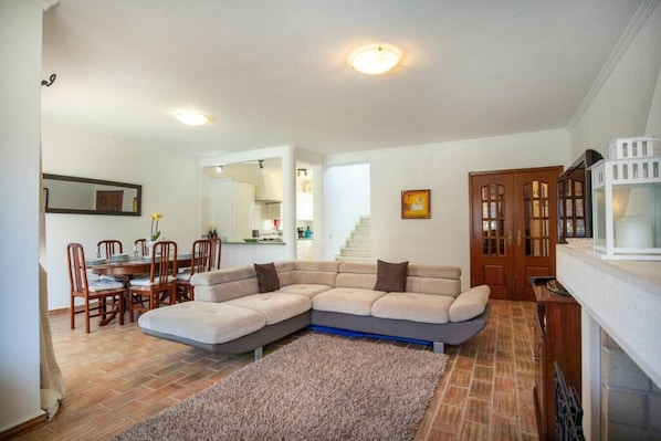 A living room with a sectional sofa, dining table, chairs, and a kitchen in the background.