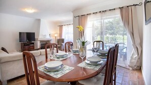 A dining area with a wooden table set for four, surrounded by chairs. A television is visible in the background, and there is a sofa and a chair in the adjacent living area.