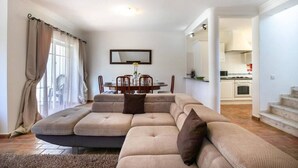 A living room with a sectional sofa, a dining table with chairs, and a kitchen in the background.