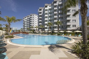 A swimming pool with lounge chairs and umbrellas, surrounded by palm trees and a multi-story building.