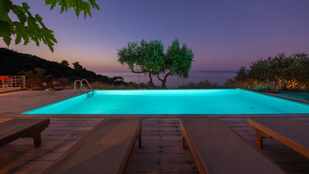 A swimming pool with wooden deck loungers, a tree in the background, and a view of the sea at dusk. - Villa Avgi (Sunrise & Pool Sea View Area) (Skopelos)