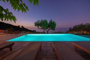 A swimming pool with wooden deck loungers, a tree in the background, and a view of the sea at dusk. - Villa Avgi (Sunrise & Pool Sea View Area) (Skopelos)