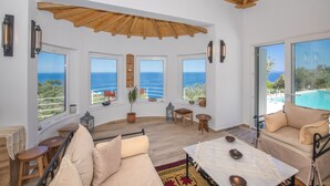 A spacious living area with large windows offering ocean views, a cozy seating area, and a wooden ceiling. - Villa Avgi (Sunrise & Pool Sea View Area) (Skopelos)