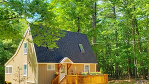 Eine zweistöckige Holzhütte mit einem dunklen Schindeldach, einer Veranda mit Holzgeländer und einer kleinen Terrassenfläche mit Blumentöpfen.