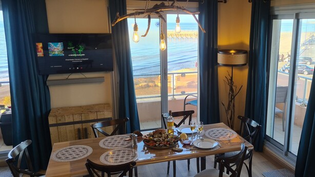 A dining area with a table set for two, a television mounted on the wall, and a view of the beach through large windows. - Le Panoramic - 180° sea view & port - Balcony - Elevator - Garage - Waterfront (Grandcamp-Maisy)