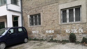 Un bâtiment à la façade en briques, doté de deux fenêtres avec des barreaux en métal, et une voiture garée devant.