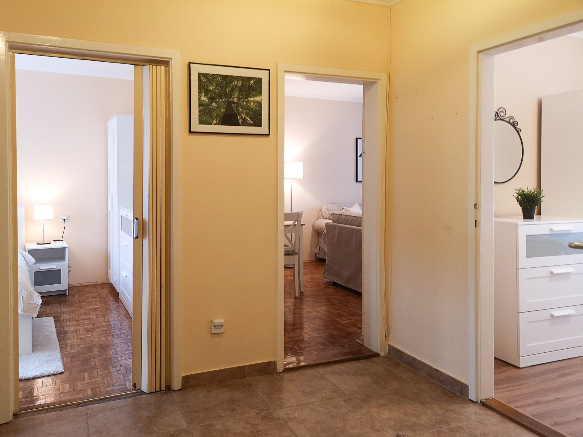 A hallway with a wooden floor, a white door, and a small white nightstand with a lamp. There is a framed picture on the wall and a potted plant on a cabinet.