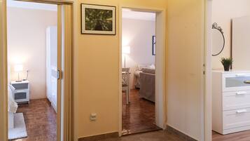 A hallway with a wooden floor, a white door, and a small white nightstand with a lamp. There is a framed picture on the wall and a potted plant on a cabinet.