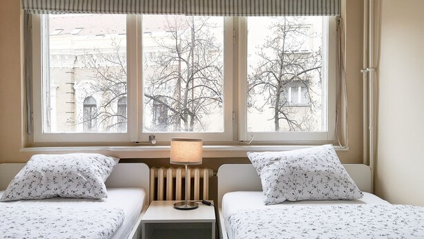 Une chambre d’hôtel avec deux lits simples, une table de chevet avec une lampe, un radiateur et une fenêtre avec un store à rayures.