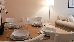 Une table à manger est dressée avec de l’assiette blanche, des verres et des couverts, entourée de chaises blanches. Une lampe sur pied assure l’éclairage, et un tableau encadré est accroché au mur.