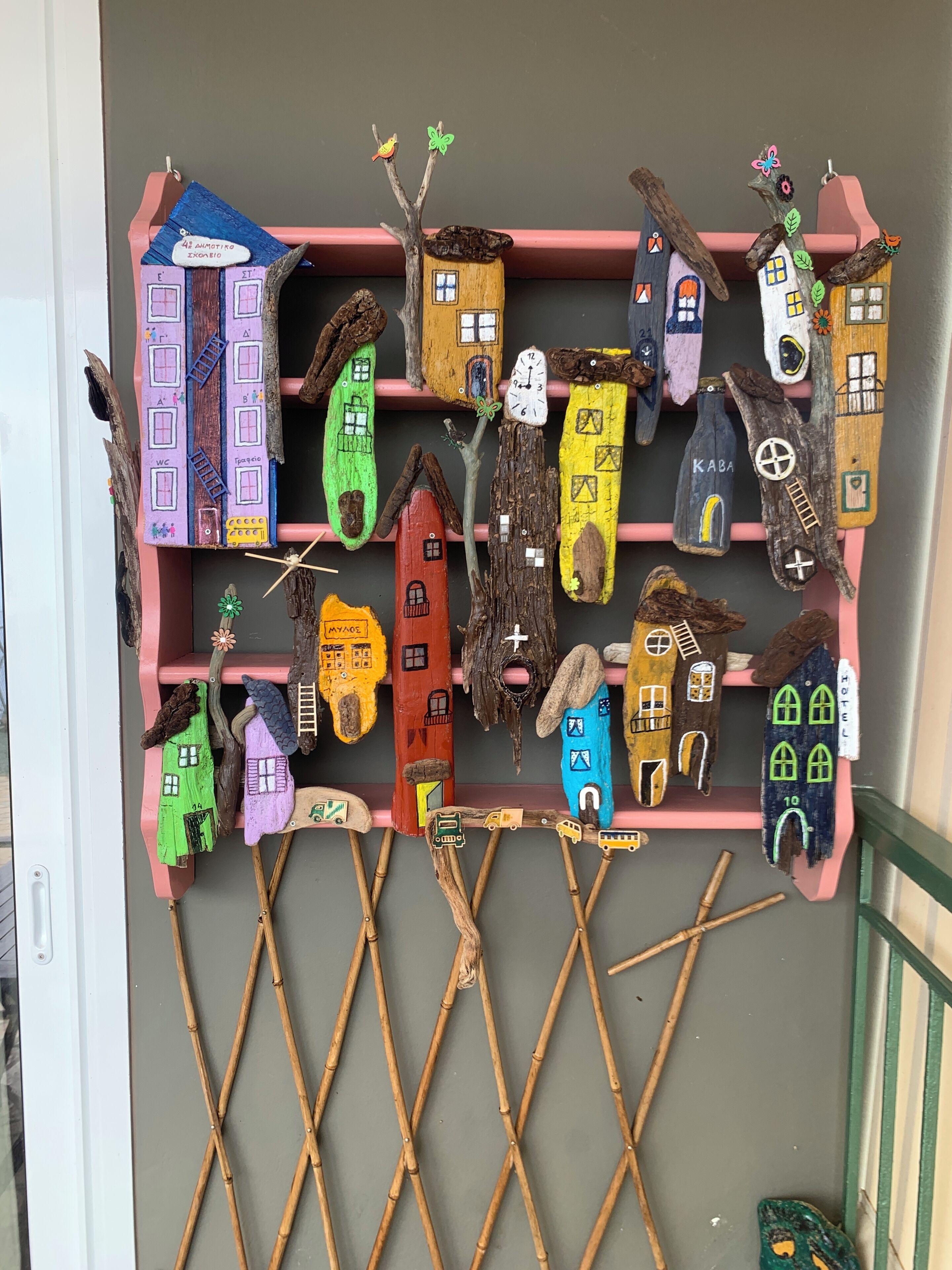 Eine Wanddekoration mit unterschiedlich farbigen und einzigartig gestalteten häuschenförmigen Objekten, manche mit Fenstern und andere mit Türen, angeordnet auf rosafarbenen Regalen vor einer grauen Wand.