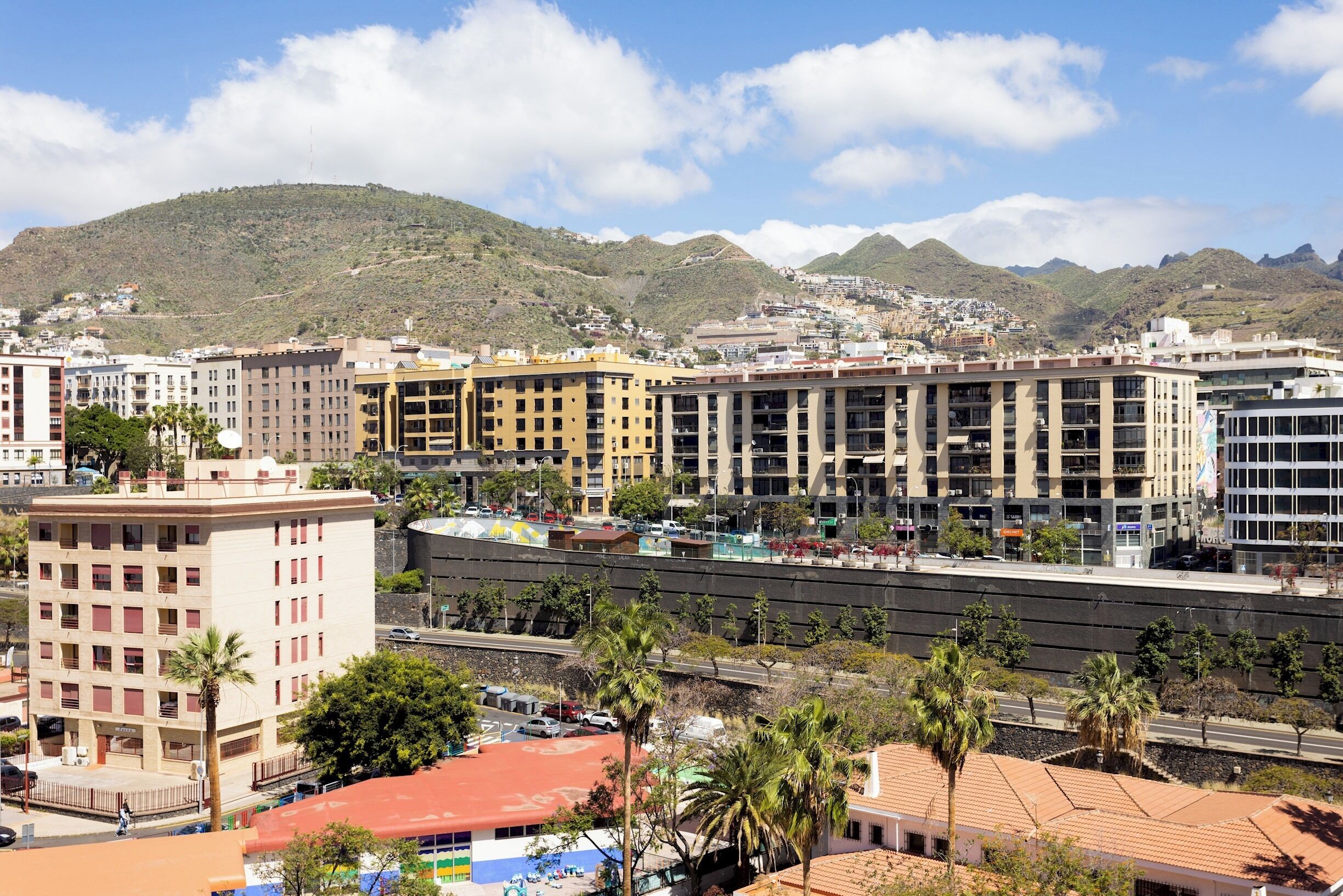 Un paysage urbain avec des bâtiments modernes, un hôtel imposant doté d’un toit rouge, et une chaîne de montagnes en arrière-plan.