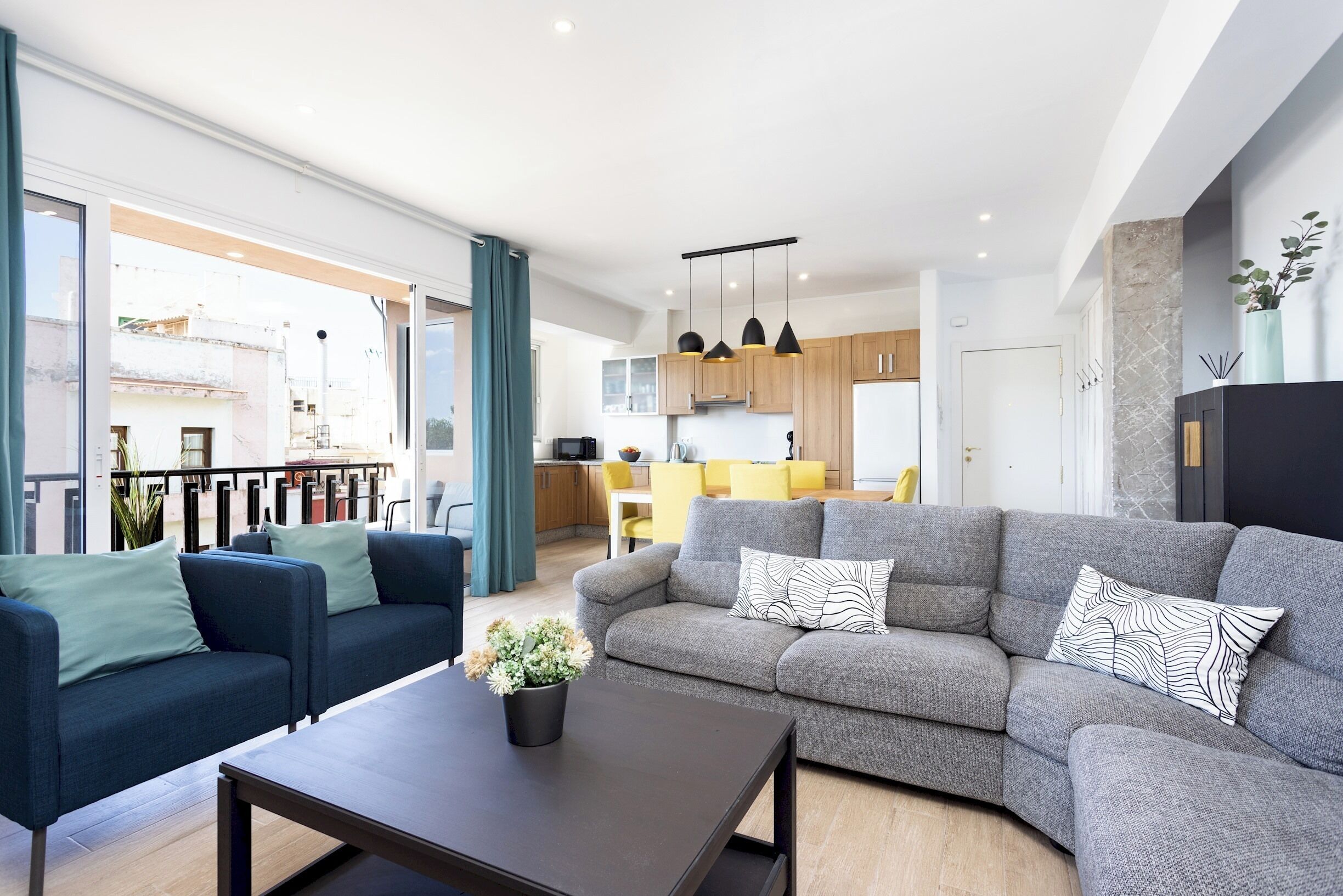 Een moderne woonkamer met een grijze bank, een blauwe fauteuil en een salontafel met een plant. Er is een keuken in de achtergrond te zien met gele stoelen.