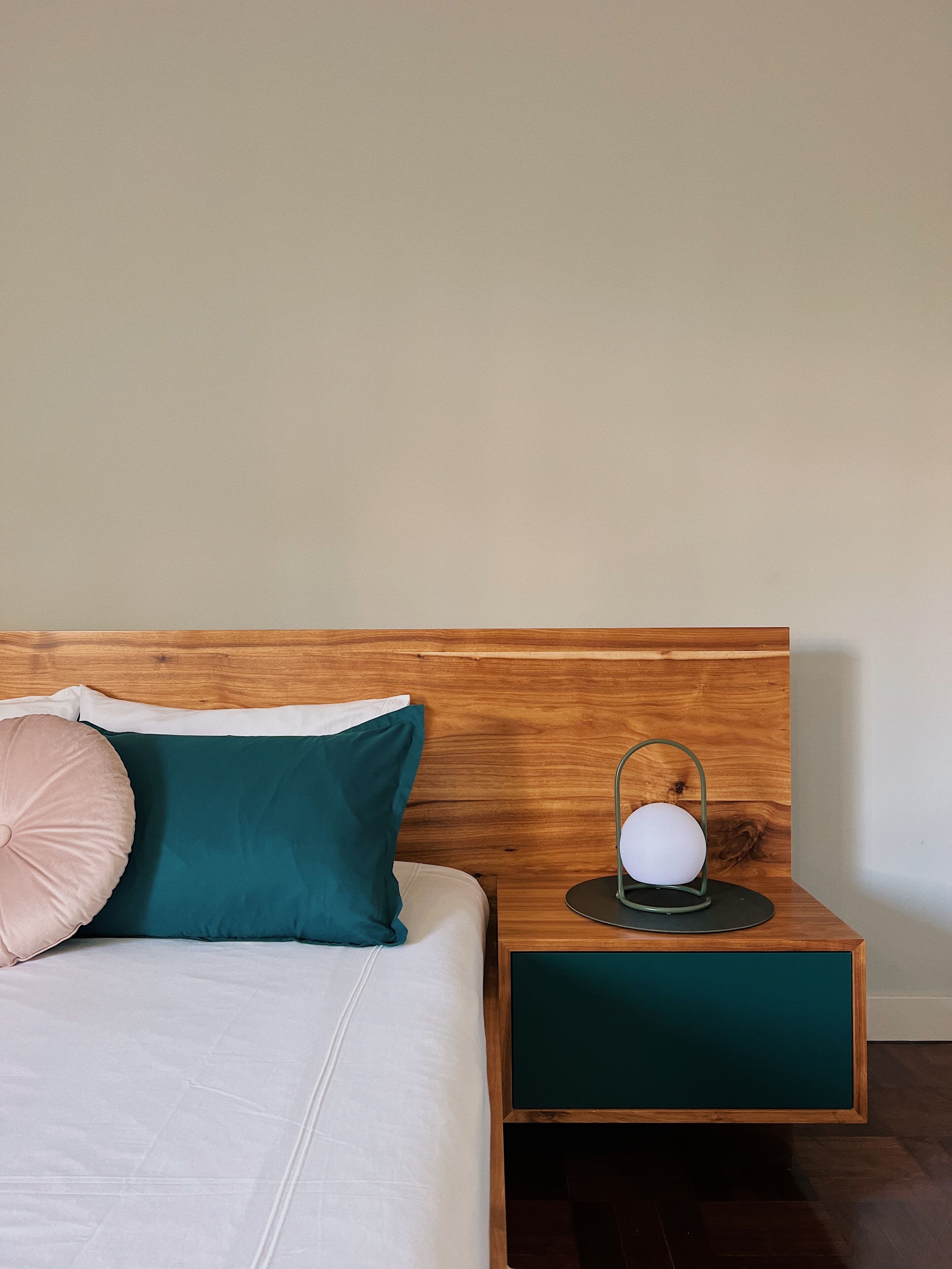 Ein ordentlich bezogenes Bett mit Holz-Kopfteil, einem grünen und einem weißen Kopfkissen. Ein Nachttisch mit grüner Schublade und einer runden Lampe darauf.
