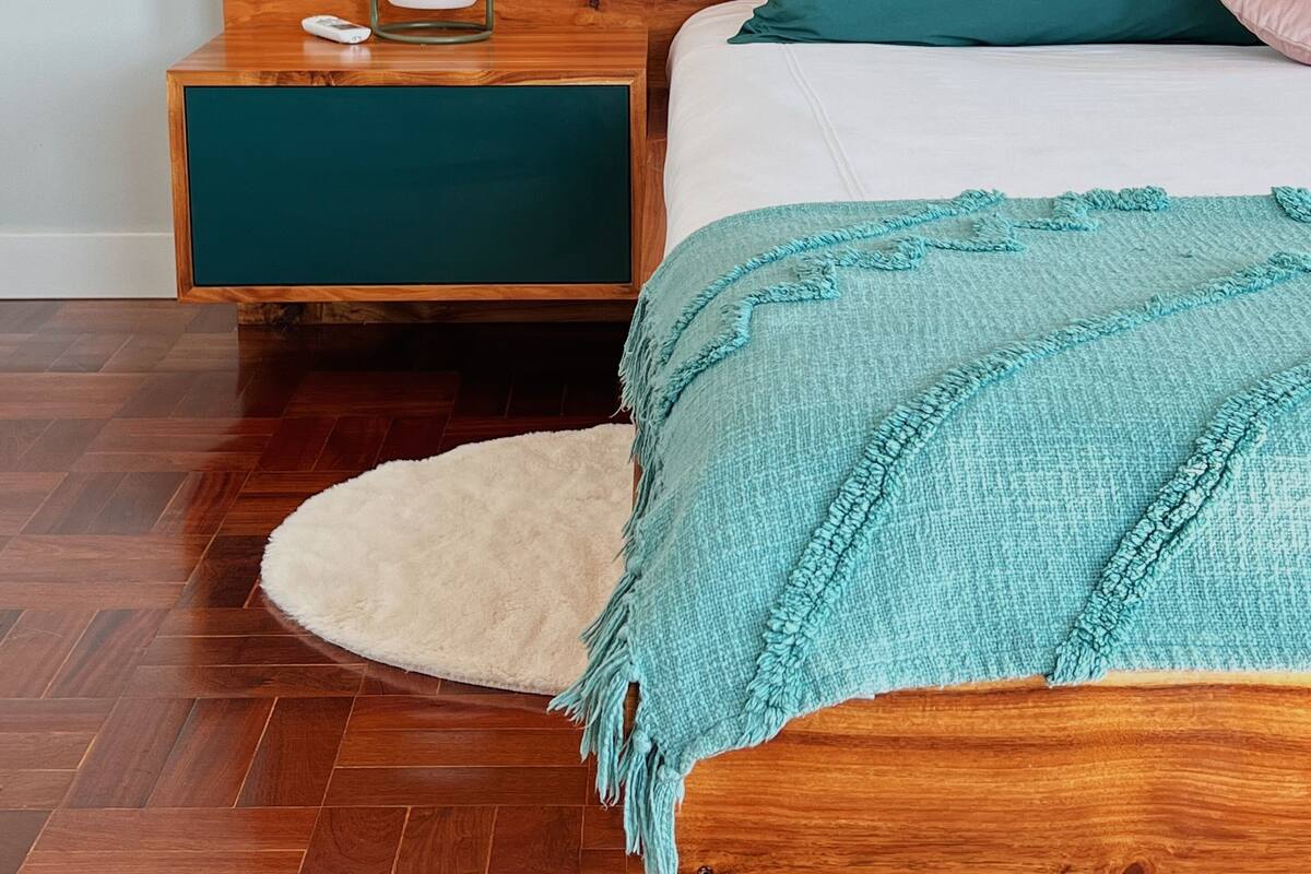 A bedroom with a wooden headboard, a white mattress, teal and pink pillows, a teal throw blanket, a white fluffy rug, and a bedside table with a lamp and a remote control.