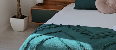 A neatly made bed with a wooden headboard, a teal throw blanket, and two pillows. A potted plant is beside the bed, and a bedside table with a lamp is visible.