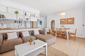 A modern living room with a sofa, coffee table, dining table, and chairs. There is a kitchen area with white cabinets and a sink.