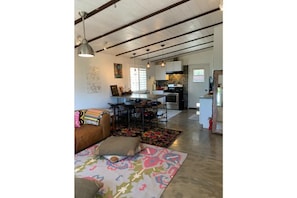 A spacious living area with a brown leather sofa, colorful rugs, and pendant lighting. A kitchen with modern appliances and a dining area with a table and chairs are visible in the background.