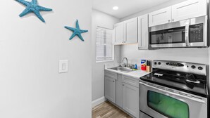 Eine moderne Küche mit weißen Schränken, einer Edelstahl-Mikrowelle und einem Backofen. An der Wand befinden sich zwei blaue Seestern-Dekorationen und ein Fenster mit Jalousien.