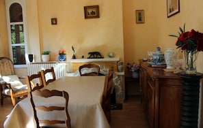 Un coin repas avec une table dressée pour quatre personnes, entourée de chaises en bois. On y trouve une cheminée dont le manteau est décoré de divers objets, notamment une figurine d’ours et un vase rempli de fleurs.