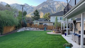 Un patio trasero con un césped cuidado, una zona de patio con mobiliario exterior y montañas al fondo.