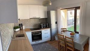 Eine kompakte Küche mit Spüle, Mikrowelle, Backofen und Kühlschrank. Ein Esstisch mit Stühlen bietet Blick auf den Garten durch das Fenster.