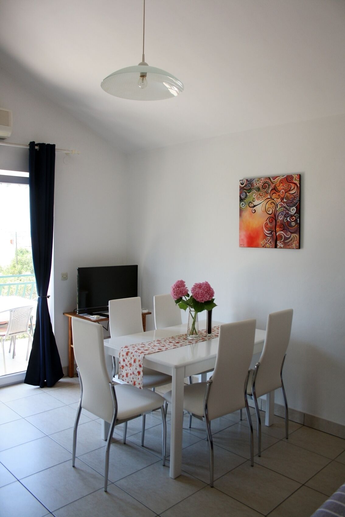 A dining area with a white table, four chairs, a vase with pink flowers, a painting on the wall, and a television on a stand.