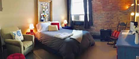Une chambre d’hôtel confortable avec un grand lit, une chaise, un petit tabouret, un bureau et une fenêtre avec des rideaux.
