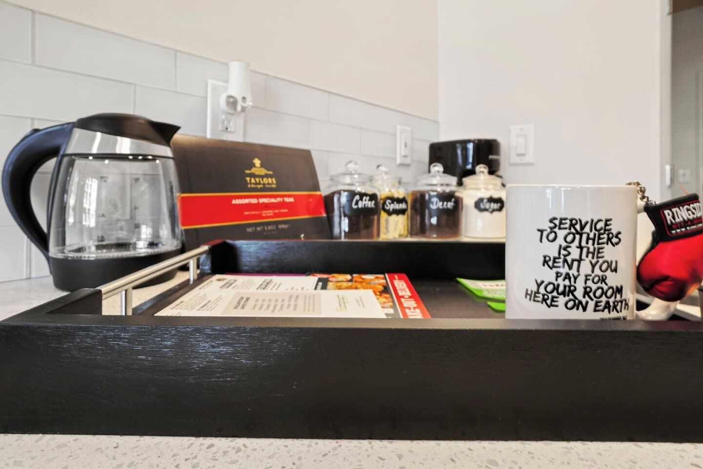 Una encimera de cocina con una cafetera, bolsitas de té y una taza con un mensaje sobre el servicio.