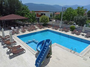 Una piscina con sillas de descanso y un tobogán, rodeada de árboles y edificios.