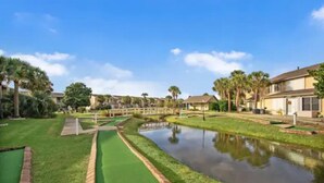 A golf course with a pond, palm trees, and houses in the background. - Cozy 2-bedroom stay with great amenities (Panama City Beach)