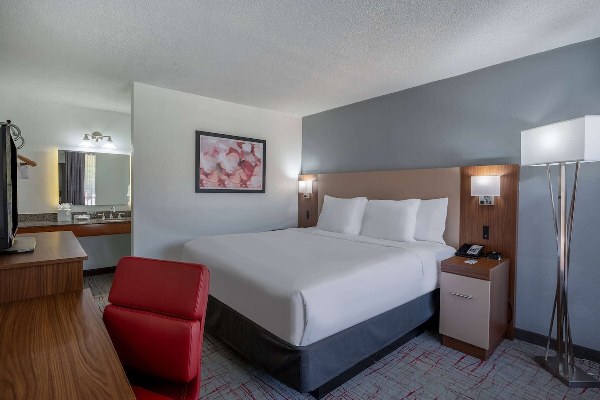 A hotel room with a large bed, a desk, a chair, a nightstand, a mirror, and a painting on the wall.