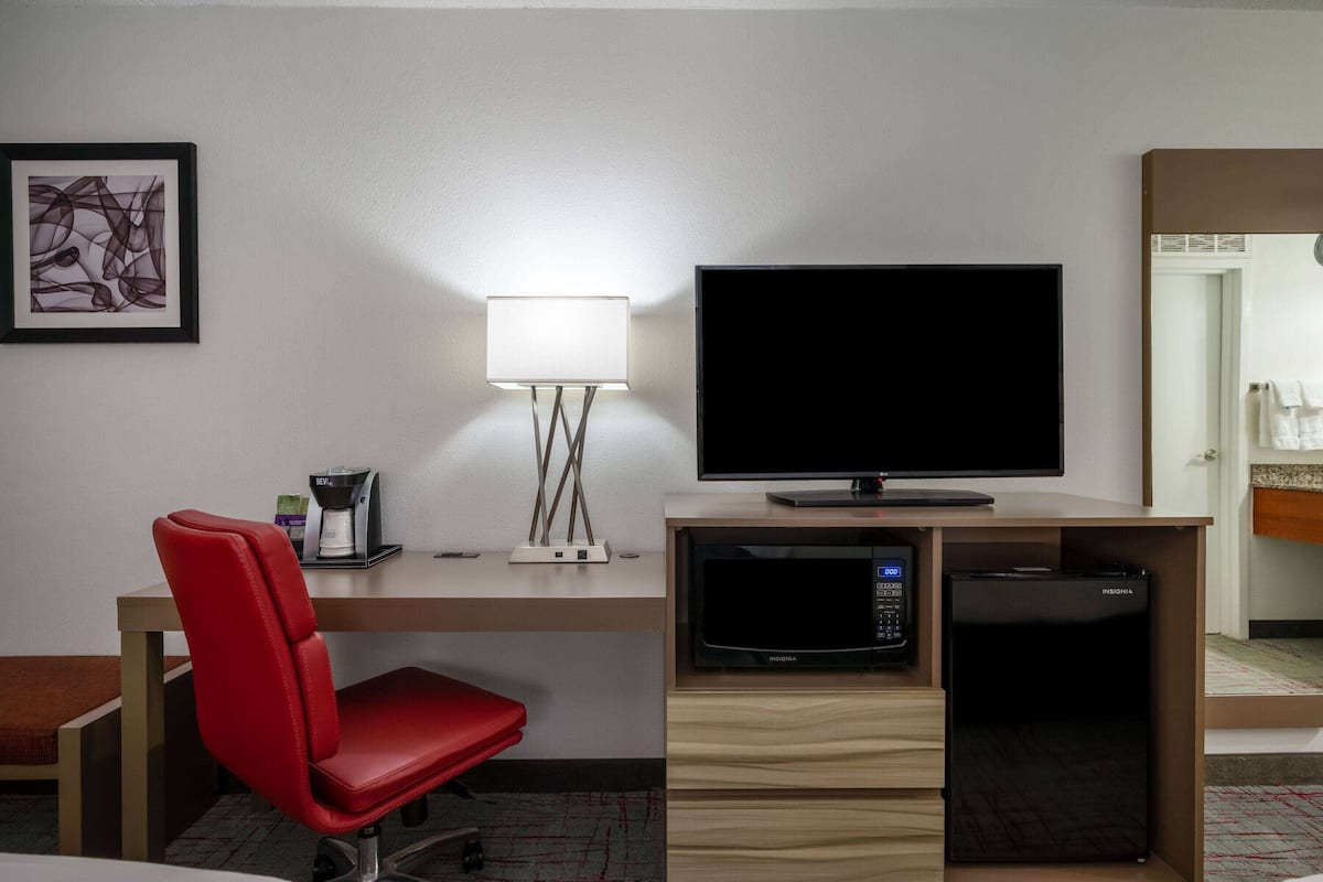 A hotel room with a desk, a red chair, a flat-screen TV, a coffee maker, a microwave, and a refrigerator.