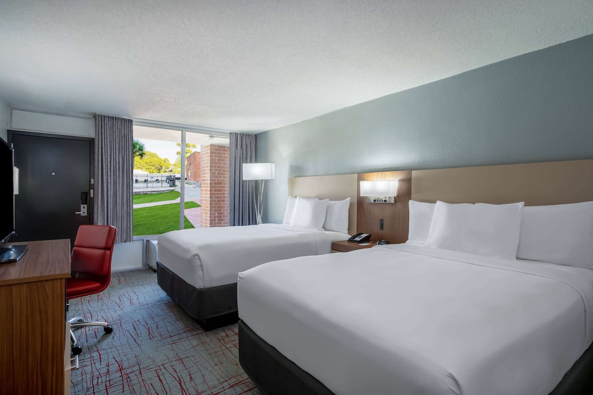 A hotel room with two beds, a desk, a chair, and a view of the outdoors.