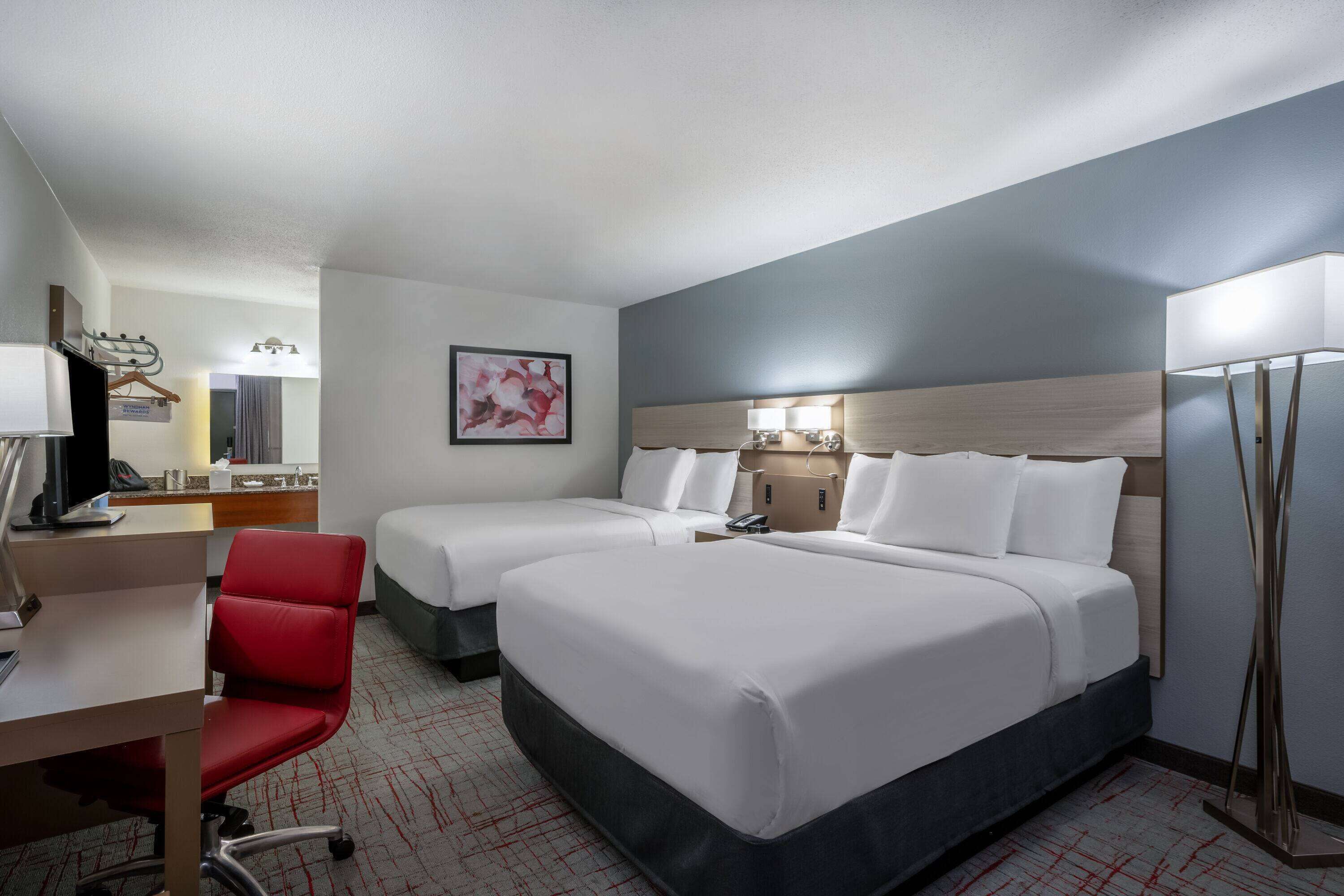 A hotel room with two beds, a desk, a chair, a desk lamp, a bedside table, and a wall-mounted TV.