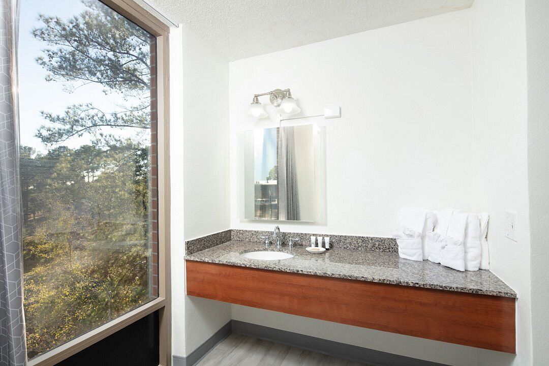 A bathroom with a large window, a granite countertop, a sink, and a mirror with lights above it.
