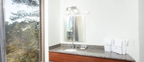 A bathroom with a large window, a granite countertop, a sink, and a mirror with lights above it.