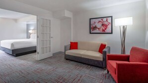 A hotel room with a bed, a gray sofa with red cushions, a red armchair, a floor lamp, and a framed abstract painting on the wall.