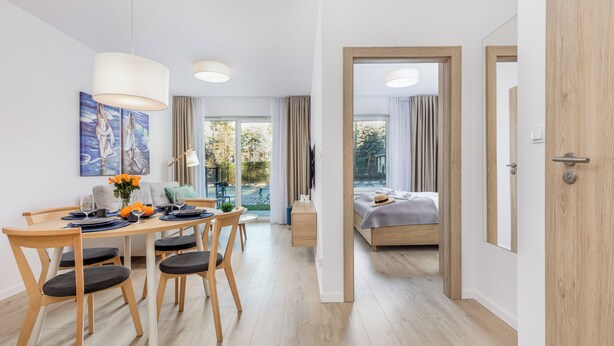 A modern hotel room with a wooden floor, a dining area with a table and chairs, a bed with a gray blanket, and a large window with curtains. - Seaside Apartamenty-Mielno  (Mielno)