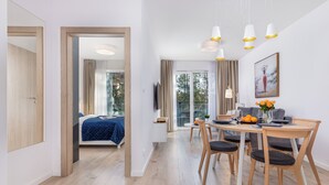 A modern hotel room with a wooden floor, a bed with a blue cover, a dining area with a table and chairs, and a large window with curtains. - Seaside Apartamenty-Mielno  (Mielno)