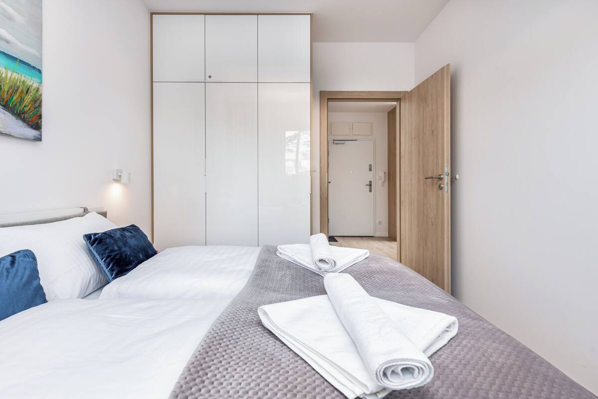 A neatly made bed with white and blue pillows, a grey textured blanket, and rolled white towels. A large wardrobe with sliding doors is visible in the background.