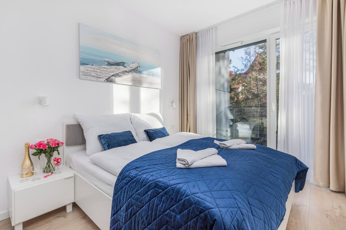 A modern bedroom with a large bed, a nightstand with a champagne bottle and glasses, and a painting of a pier on the wall.