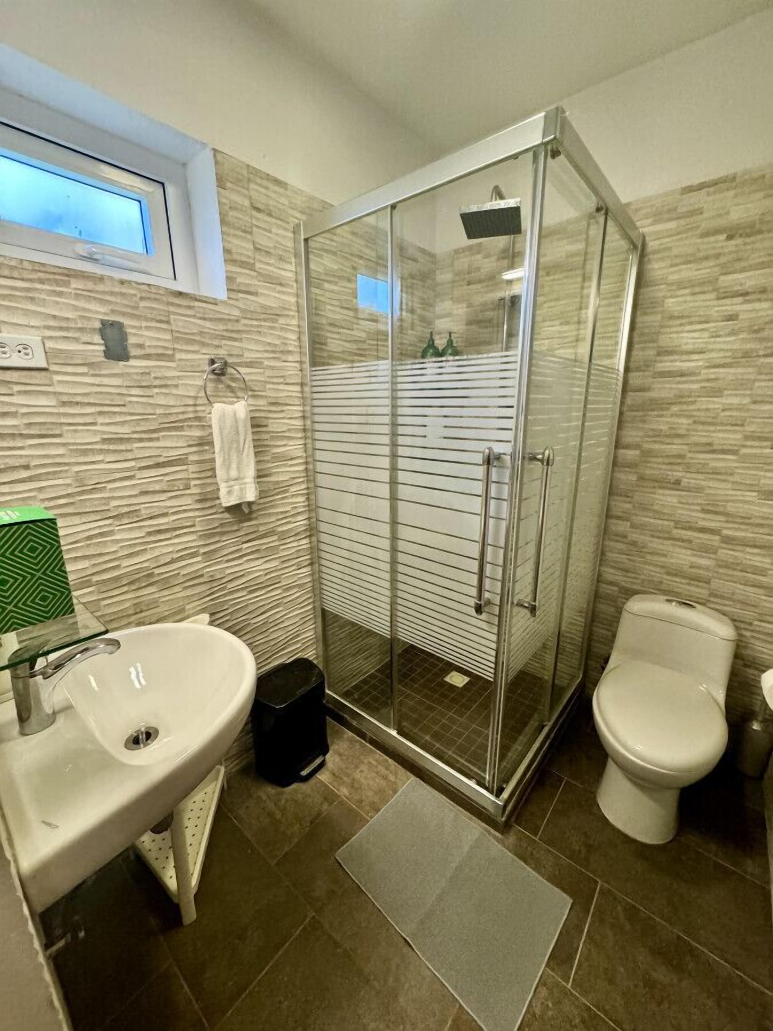 Ett modernt badrum med en dusch som är avskärmad med glasväggar, ett handfat med en grön tvålbehållare och en toalett.