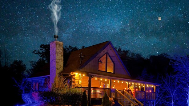A house with a chimney, a porch with string lights, and a visible moon in the night sky. - Bootlegger Cabin | Hot Tub | NERF Wall | Arcade (Sevierville)