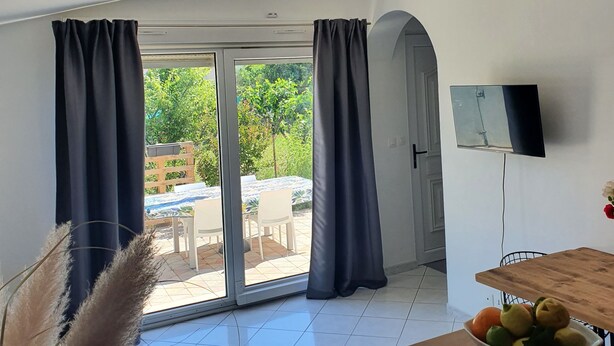 A room with a large sliding glass door leading to an outdoor area, a flat-screen TV mounted on the wall, and a wooden table with a bowl of fruit. - Apartment (Caux)