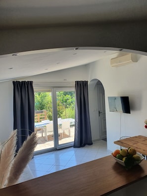Quarto com uma grande porta de vidro deslizante que dá para uma área externa, uma televisão de tela plana na parede e uma mesa de madeira com uma tigela de frutas.