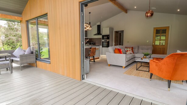 A modern interior with a wooden exterior, a dining area with a glass door, a living room with a grey sofa and orange armchair, and a kitchen in the background. - Pool View (Meifod)
